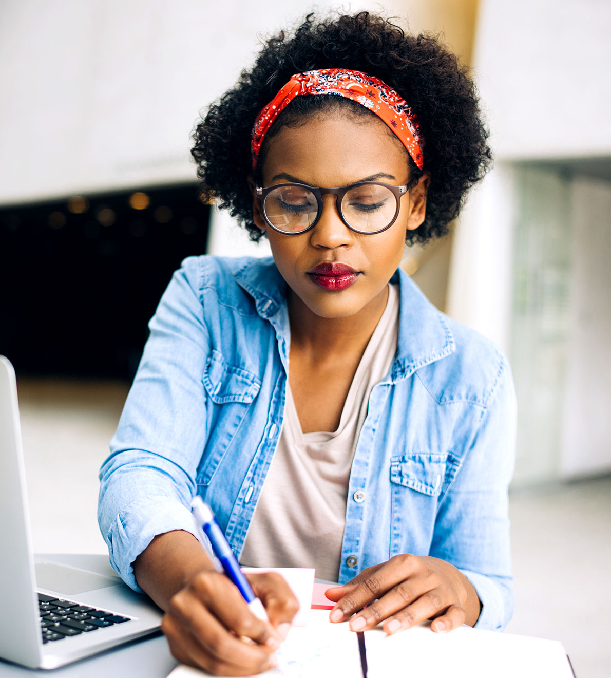 info-5 Woman in glasses and a denim shirt taking notes beside her laptop